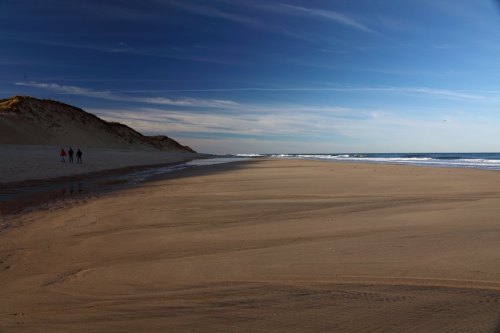 A serene beach scene with gentle waves, sandy shore, and three people walking along the water's edge under a blue sky.