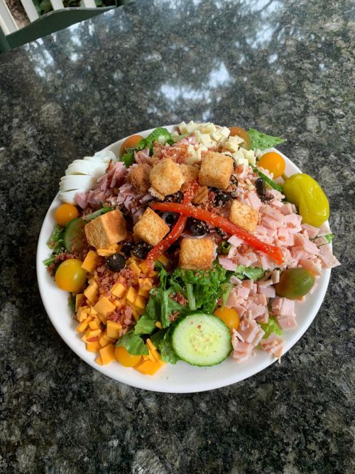 A colorful salad with greens, diced ham, cheese, cherry tomatoes, olives, croutons, and peppers on a white plate.