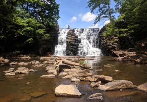 A serene waterfall cascades over rocks, surrounded by lush greenery and a calm river with scattered stones in the foreground.