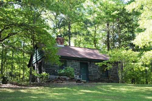 A small stone cabin surrounded by lush green trees and grass, featuring a chimney and a simple front porch.