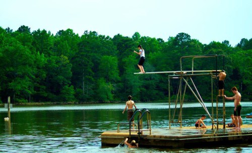 A group of kids enjoying a lake, with one jumping off a diving platform and others swimming and playing nearby.