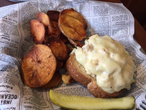 A plate featuring a sandwich with creamy topping, sweet potato chips, and a pickle on newspaper-style wrapping.