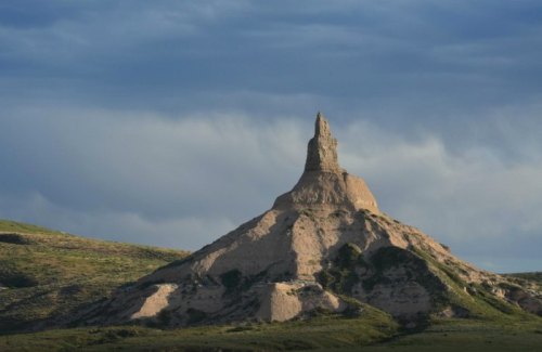 A tall, rocky formation rises from a grassy landscape under a cloudy sky.