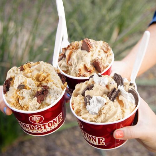 Three hands hold large cups of ice cream, each topped with various mix-ins like cookies and chocolate.