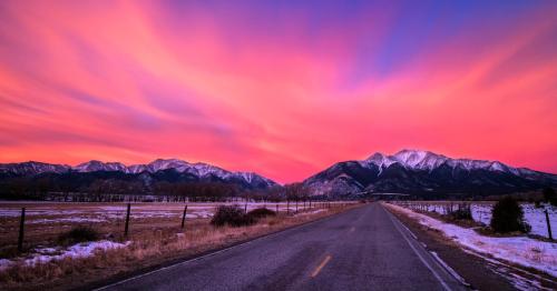 A scenic road leads towards majestic mountains under a vibrant pink and purple sunset sky. Snow covers the ground.