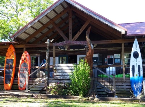 A rustic wooden building with kayaks and paddleboards outside, surrounded by greenery.
