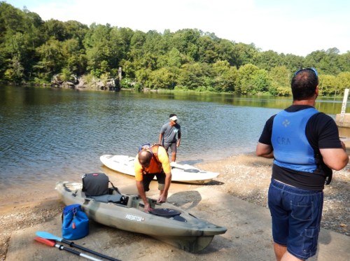 Three people prepare kayaks by a calm lake surrounded by green trees on a sunny day.