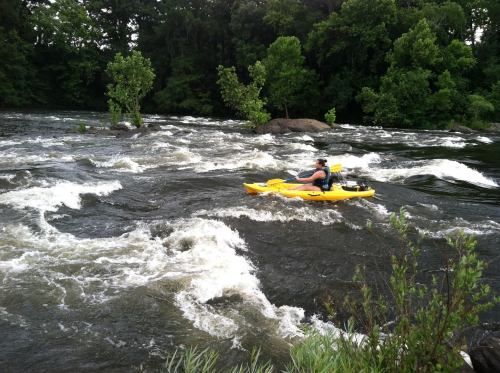 A person paddles a yellow kayak through turbulent river rapids surrounded by lush green trees.