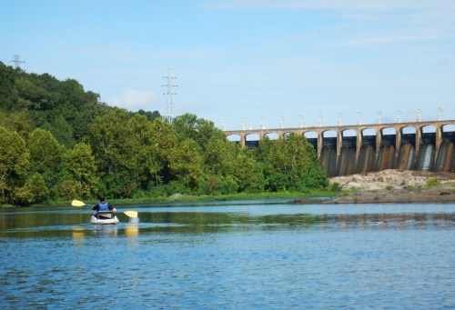 A person kayaking on a calm river with a dam and lush greenery in the background under a clear blue sky.