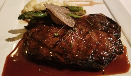 A beautifully plated steak with asparagus and mashed potatoes, garnished with truffle slices and herbs.