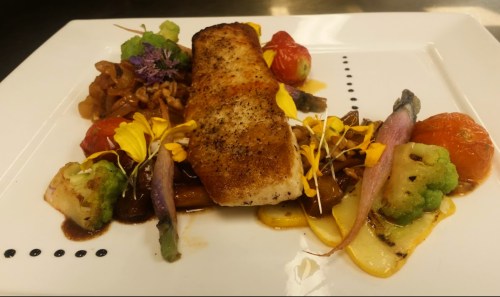 A beautifully plated dish featuring seared fish, colorful vegetables, and garnishes on a white plate.