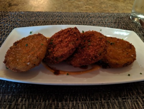 Four golden-brown fried green tomato slices on a white plate, garnished with green herbs.