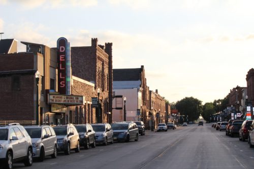 A quiet street lined with parked cars and historic brick buildings, featuring a marquee sign for "Dells."