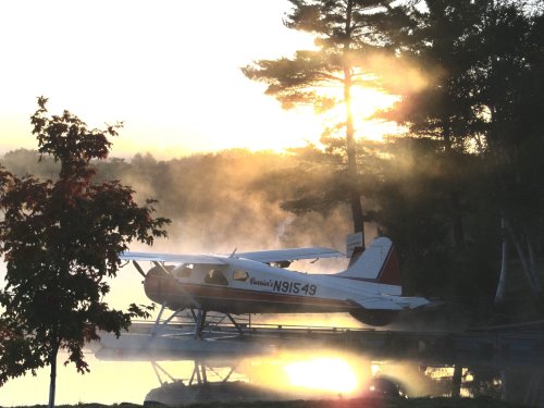 A seaplane sits on a misty lake at sunrise, surrounded by trees and soft golden light.
