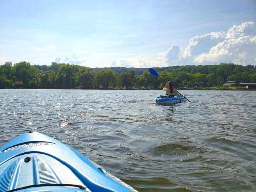 A person kayaking on a calm lake with lush green trees and blue skies in the background.
