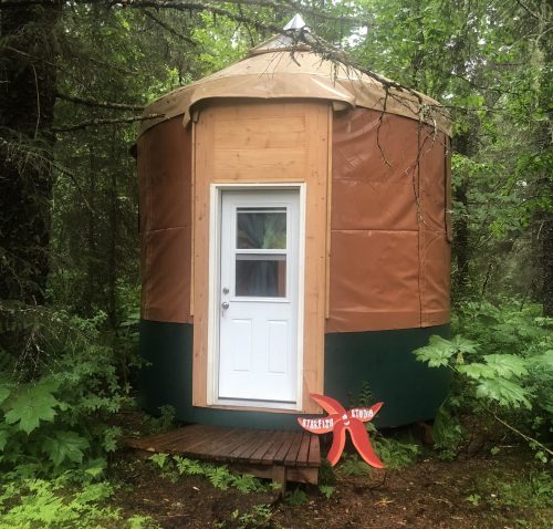 A small yurt with a wooden door, surrounded by lush greenery and trees, sits on a wooden platform.