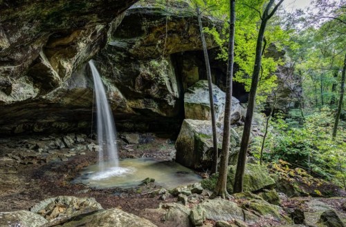 A serene waterfall cascades from a rocky overhang into a small pool, surrounded by lush green trees and boulders.