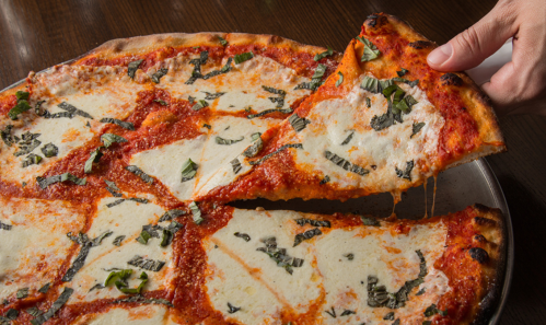 A hand lifts a slice of cheese pizza topped with basil from a whole pizza on a wooden table.