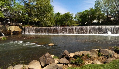 A serene landscape featuring a waterfall cascading over a stone dam, surrounded by lush greenery and a clear blue sky.