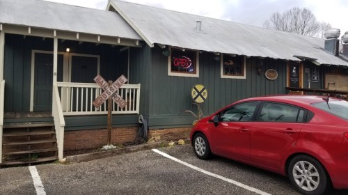 A red car parked outside a rustic green building with a metal roof and a neon "open" sign.