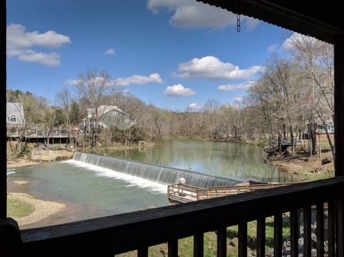 A serene view of a river with a small waterfall, surrounded by trees and houses under a blue sky with fluffy clouds.
