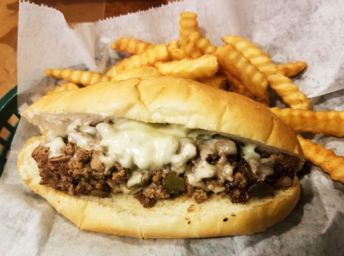 A close-up of a cheesesteak sandwich with melted cheese, served with crinkle-cut fries on the side.