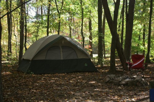 A tent set up in a wooded area with autumn leaves, alongside a red camping chair and a stone fire pit.