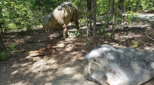 A dinosaur statue in a wooded area, with a nearby rock displaying the name "Coelophysis."