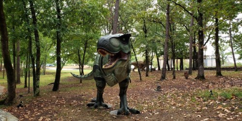 A life-sized dinosaur statue stands among trees in a park, surrounded by fallen leaves and greenery.