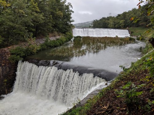 A scenic view of a waterfall cascading over a dam, surrounded by lush greenery and misty hills in the background.