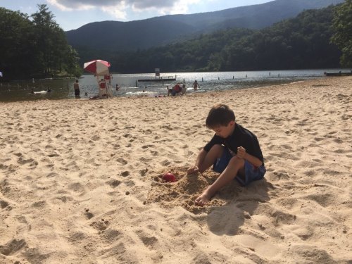 A child playing in the sand at a beach near a lake, with mountains in the background and people enjoying the water.