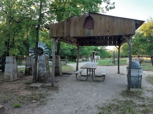 A rustic picnic area with a wooden shelter, picnic table, and a windmill surrounded by trees and gravel paths.