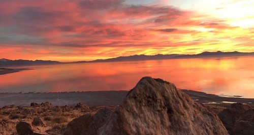 A vibrant sunset over a calm lake, with colorful clouds reflecting on the water and rocky foreground.