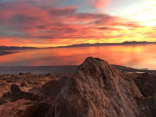A vibrant sunset over a calm lake, with colorful clouds reflecting on the water and rocky foreground.