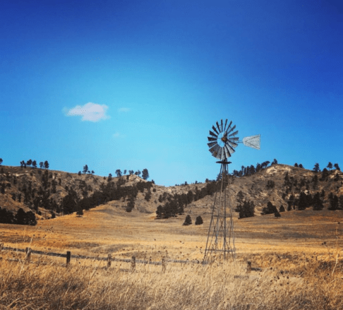 A windmill stands in a grassy field with rolling hills and a clear blue sky in the background.
