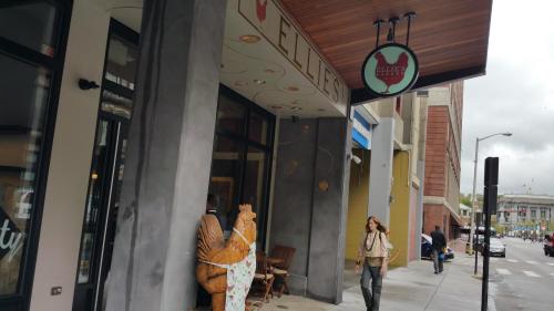 A woman walks past a café entrance with a large chicken sculpture outside, on a cloudy day in an urban setting.