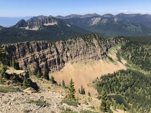 A panoramic view of rugged mountains and a valley, featuring steep cliffs and lush greenery under a clear blue sky.