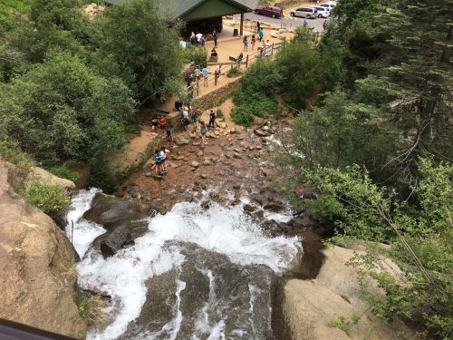 A waterfall cascades down rocks, with people exploring a path and greenery surrounding the area.