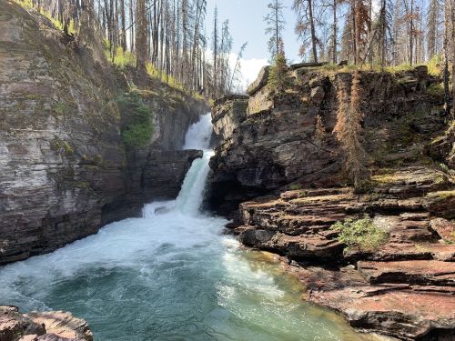A waterfall cascades into a turquoise pool, surrounded by rocky cliffs and sparse trees in a sunlit landscape.