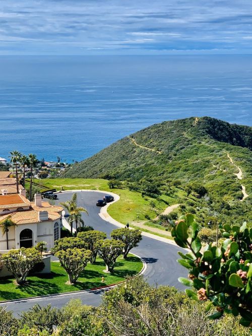 A scenic view of a coastal landscape with rolling hills, a winding road, and a glimpse of the ocean under a cloudy sky.