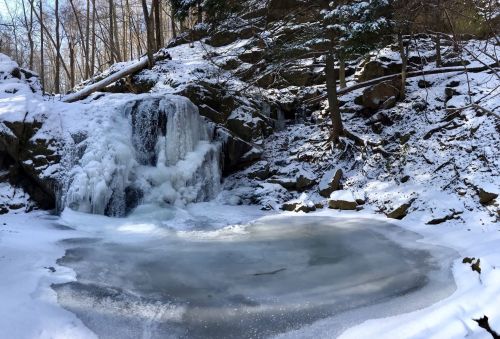 A frozen waterfall surrounded by snow-covered rocks and trees, with a partially frozen pond in the foreground.