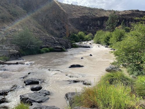 A winding river flows through rocky terrain, surrounded by lush greenery and hills under a cloudy sky.