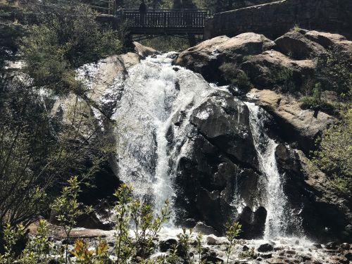 A waterfall cascades over rocky terrain, surrounded by greenery, with a bridge visible in the background.