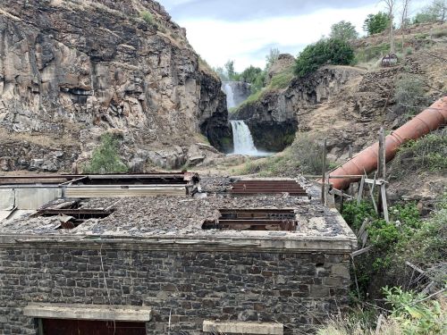 An abandoned building with a crumbling roof in the foreground, with a waterfall cascading down rocky cliffs in the background.