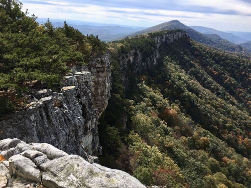 A scenic view of a rocky cliff overlooking a lush, forested valley and distant mountains under a cloudy sky.