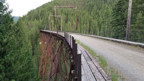 A rusted metal bridge spans a forested valley, with a gravel path running alongside it amidst tall green trees.
