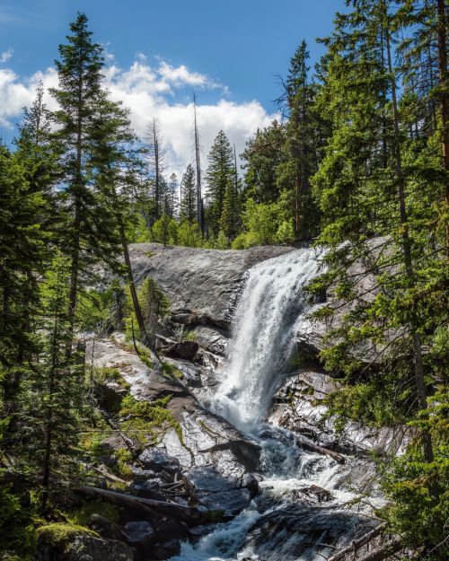 A serene waterfall cascades over rocky terrain, surrounded by lush green trees and a bright blue sky.