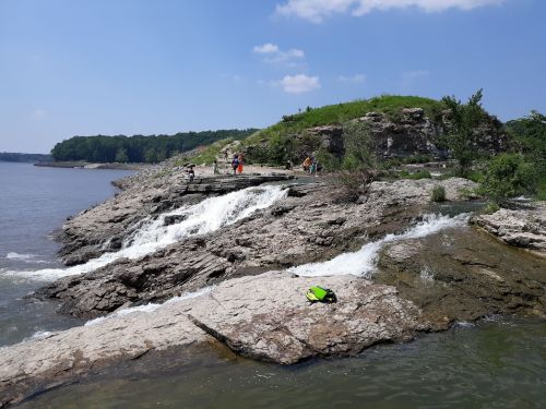 A rocky shoreline with gentle waves, people exploring the area, and greenery in the background under a blue sky.