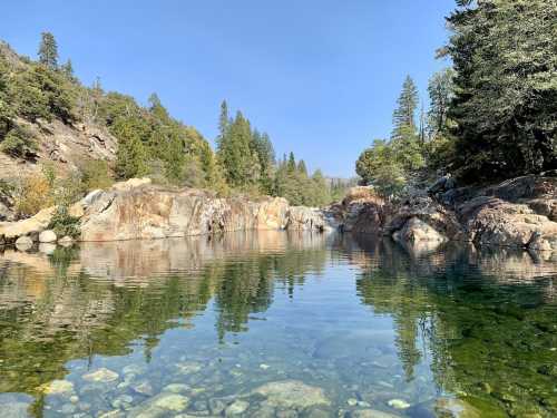 A serene river scene with clear water reflecting trees and rocky banks under a blue sky.