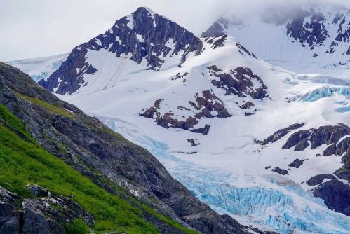 Snow-capped mountains with a glacier and green slopes in the foreground under a cloudy sky.